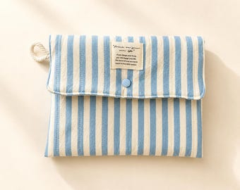 May include: A rectangular fabric pouch featuring vertical light blue and cream stripes. The flap includes a small light blue button and a rectangular white label with the text "Focus on your own life" and additional smaller printed text.