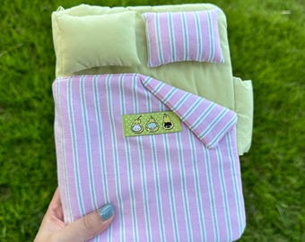May include: A miniature bed set featuring a light green fitted sheet and a matching solid light green pillow. The bedding includes a pink and white striped duvet with a rectangular patch displaying three illustrated pear-shaped characters.