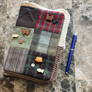 Op de afbeelding: Een patchwork stoffen etui met een verscheidenheid aan texturen en patronen, waaronder visgraat en ruitjes. De etui is versierd met houten knopen in de vorm van dieren en een witte gebreide rand. Een blauwe pen ligt ernaast.