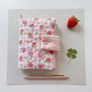 May include: A pink quilted notebook with a strawberry pattern, a small matching pocket, and a rose gold pen. A fresh strawberry and a four-leaf clover are also present. The notebook has a gingham pattern and a heart-shaped clasp.