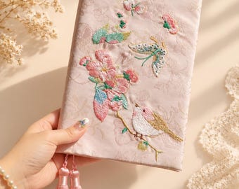 May include: A light pink fabric-covered journal featuring intricate embroidery of a bird perched on a flowering branch, accompanied by butterflies. The cover has a subtle tonal floral pattern and two matching pink tassels hanging from the bottom.
