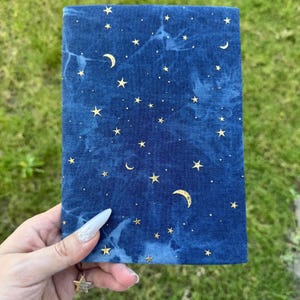 Könnte beinhalten: Ein rechteckiges, marineblaues Notizbuch mit einem Himmelsdesign. Der Einband zeigt goldene Sterne und Halbmonde auf einem marmorierten, dunkelblauen Hintergrund. Das Notizbuch wird vor einem Hintergrund aus grünem Gras gehalten.