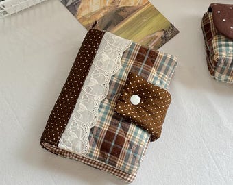 May include: A quilted fabric wallet featuring a brown and white polka dot pattern on the spine and closure flap. The main body has a brown, blue, and cream plaid design accented by a white lace trim with heart-shaped cutouts and a white button.