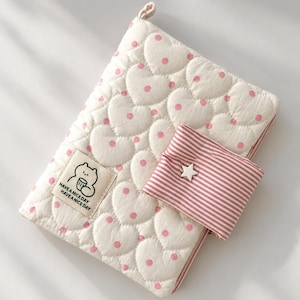 May include: A quilted off-white fabric cover featuring a repeating heart pattern with small pink dots. A rectangular patch displays a cartoon bear holding a cup and the text HAVE A NICE DAY. A pink and white striped flap features a white star button.