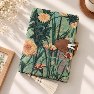 May include: A rectangular journal with a woven fabric cover featuring a green background and a floral pattern of orange and yellow dandelions. The journal includes a brown faux leather toggle closure with a white cord and a decorative cream tassel.