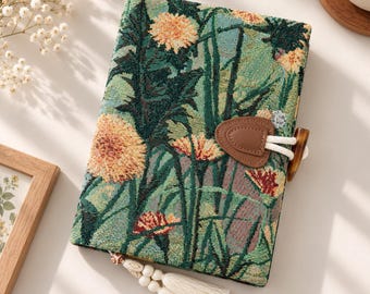 May include: A rectangular journal with a woven fabric cover featuring a green background and a floral pattern of orange and yellow dandelions. The journal includes a brown faux leather toggle closure with a white cord and a decorative cream tassel.