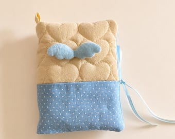 May include: A rectangular fabric pouch featuring a quilted beige upper section with a heart pattern and a pair of light blue felt wings attached. The lower section is light blue with a white polka dot pattern, finished with light blue ribbon ties.