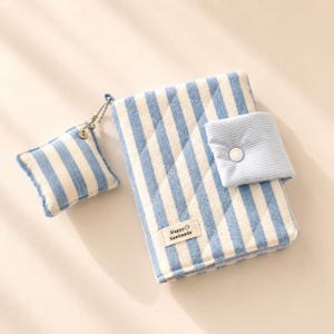 May include: A quilted fabric notebook cover featuring a blue and white vertical stripe pattern. It includes a matching square charm on a metal chain and a snap-button closure flap. A small rectangular label reads Happy handmade.