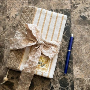 Könnte beinhalten: Ein beige-weiß gestreiftes Notizbuch mit gewebter Textur, gebunden mit einem hellrosa Spitzenband. Ein blauer Stift liegt neben dem Notizbuch. Das Notizbuch hat eine dekorative Briefmarke mit den Worten "Bonne Courage".