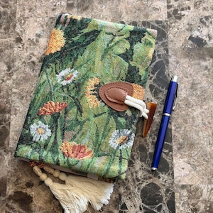 May include: A journal with a floral tapestry cover, brown leather clasp, and a blue pen. The cover displays a green backdrop with yellow, orange, and white flower patterns. A cream tassel and wooden beads are attached to the journal.