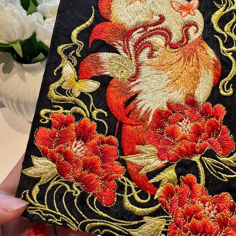 May include: Black fabric with detailed embroidery of a fox, butterflies, and flowers in red, orange, and gold. The design is ornate, with a focus on floral and animal motifs, showcasing a rich colour palette.