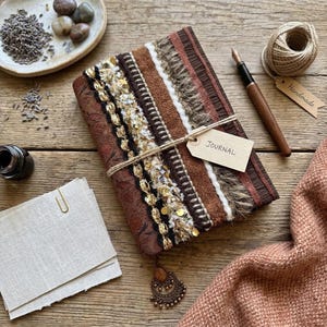 May include: A handmade journal with a decorative cover featuring brown, gold, and white textured fabrics. The journal is tied with twine and has a tag that reads "Journal". A pen, inkwell, and other stationery items are nearby.