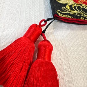 May include: A close-up shot of two bright red tassels with black cords, attached to a black fabric with gold and red embroidery. The tassels are made of many strands of red thread. The background is a white, textured surface.