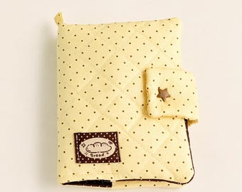 May include: A quilted light yellow notebook cover featuring a brown polka dot pattern. It includes a small tab closure with a brown star-shaped button and a rectangular brown patch with a white illustration of bread and the text Bread.