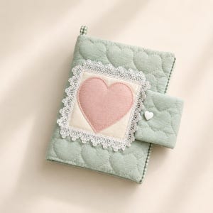 May include: A sage green quilted notebook cover featuring a repeating heart pattern. The front displays a square patch with a pink fabric heart, framed by white lace trim. A matching quilted strap with a small white heart button secures the cover.