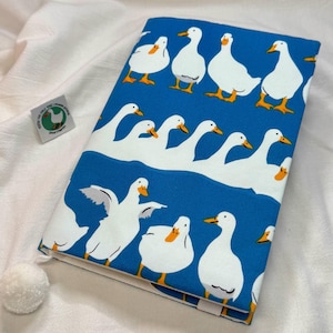 May include: A rectangular fabric book cover featuring a vibrant blue background with a repeating pattern of white ducks with orange beaks and feet. The cover includes a small white pom-pom attached to the corner and a small square tag with a duck graphic.