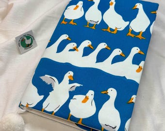 May include: A rectangular fabric book cover featuring a vibrant blue background with a repeating pattern of white ducks with orange beaks and feet. The cover includes a small white pom-pom attached to the corner and a small square tag with a duck graphic.