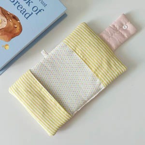May include: A rectangular fabric pouch featuring a white center panel with small multicolored polka dots, flanked by yellow and white striped side panels. A pale pink fabric flap with a single white snap closure extends from the top edge.