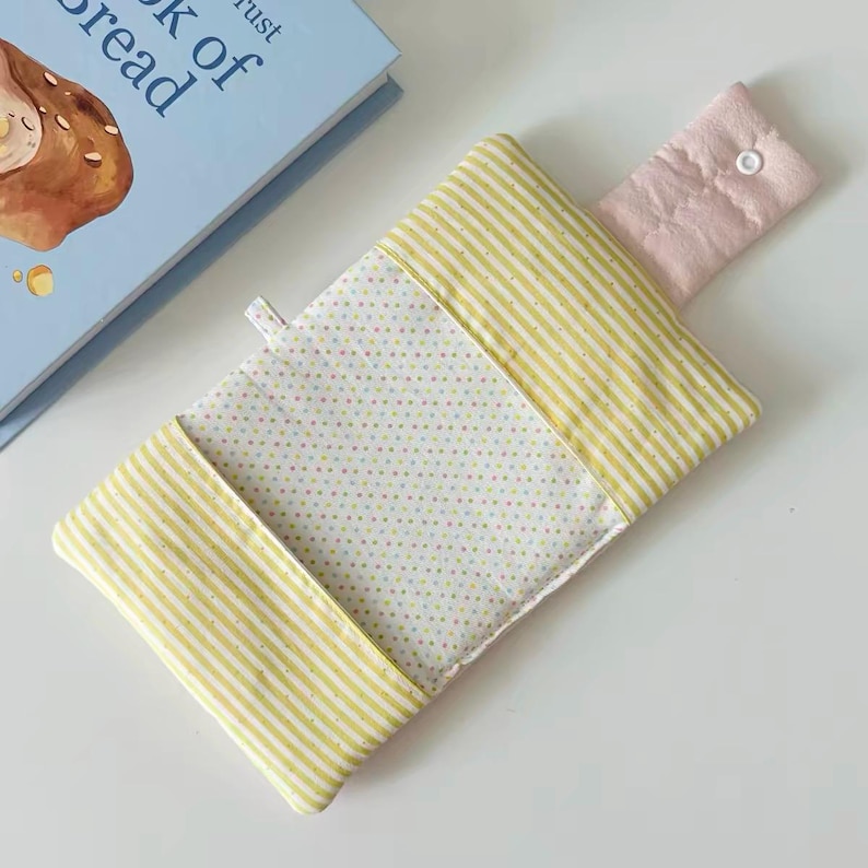 May include: A rectangular fabric pouch featuring a white center panel with small multicolored polka dots, flanked by yellow and white striped side panels. A pale pink fabric flap with a single white snap closure extends from the top edge.