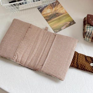 May include: A rectangular fabric pouch featuring a brown and white gingham pattern. The pouch has a fold-over flap closure made of brown fabric with white polka dots, secured by a single white snap button. The exterior is quilted.