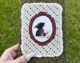 May include: A rectangular fabric-covered journal featuring an olive green floral print base. The centre panel has a cream background with black polka dots, framed by white lace trim and a dark red oval border depicting a black bird and a small mouse.