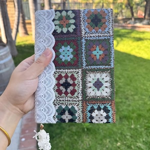 May include: A colorful, patterned journal with a white lace trim and a tassel. The journal's cover features a patchwork design with squares of various colors and floral patterns. The lace trim adds a delicate touch to the design.