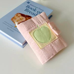 May include: A light pink quilted fabric book cover featuring a centered square patch with a light green apple appliqu&eacute;. The cover has a small fabric tab closure on the right side and is resting on top of a National Trust Book of Bread.