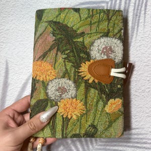 May include: A floral tapestry journal with a brown leather closure. The cover features a green and yellow floral pattern with dandelions. The journal is closed with a brown toggle and white cord.