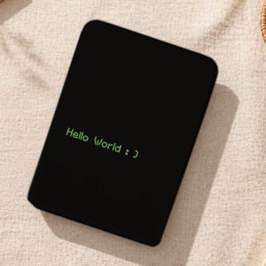 Könnte beinhalten: Ein schwarzes Tablet mit dem grünen Text "Hello World :)". Das Tablet liegt auf einer beigen Oberfläche, im Hintergrund sind ein Stift und ein gewebter Untersetzer zu sehen. Das Bild hat eine minimalistische Ästhetik.