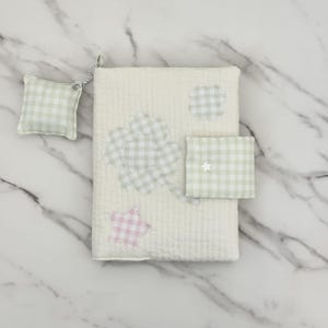 May include: A rectangular off-white quilted fabric book cover featuring a large light green gingham four-leaf clover appliqué, a small light green gingham cloud, a pink gingham star, and a light green gingham tab with a small white star.
