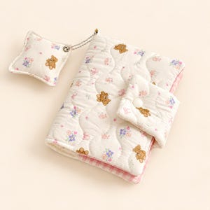 Peut inclure: Une couverture de carnet en tissu matelassé avec un motif répétitif d'oursons bruns et de petites fleurs roses et bleues sur fond blanc. La couverture comprend une fermeture à bouton-pression et une petite breloque carrée assortie attachée par une chaîne.