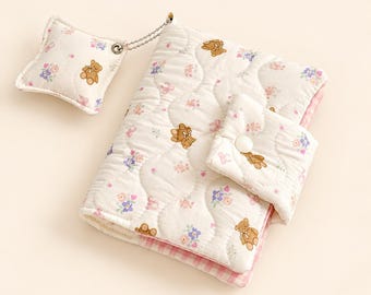 May include: A quilted fabric notebook cover featuring a repeating pattern of brown teddy bears and small pink and blue flowers on a white background. The cover includes a snap button closure and a small, square, matching quilted charm attached by a chain.