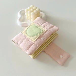 May include: A quilted pink fabric pouch featuring a light green apple appliqu&eacute; on a square patch. The pouch has a flap with a white snap button closure and reveals a yellow and white striped interior lining. A cream-colored bubble candle sits nearby.