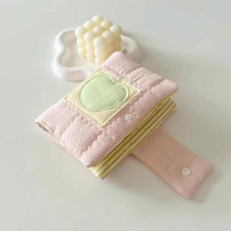 May include: A quilted pink fabric pouch featuring a light green apple appliqu&eacute; on a square patch. The pouch has a flap with a white snap button closure and reveals a yellow and white striped interior lining. A cream-colored bubble candle sits nearby.
