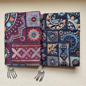 Op de afbeelding: Twee decoratieve tijdschriften met ingewikkelde borduursels in bordeauxrood en marineblauw. De tijdschriften hebben bloemen- en geometrische patronen met pailletten. Zilveren oorbellen met bungelende elementen zijn eronder geplaatst.