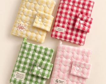 May include: Four quilted fabric notebook covers in yellow, red, green, and pink gingham patterns. Each cover features a heart-quilted texture, a small attached pocket with a white heart button, and a rectangular label with text.