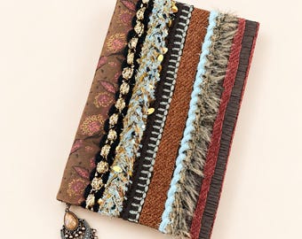 May include: A rectangular journal cover featuring a brown paisley fabric panel accented by vertical strips of black braided trim, light blue textured yarn, silver metallic stitching, brown woven fabric, and a light blue twisted cord with fringe.