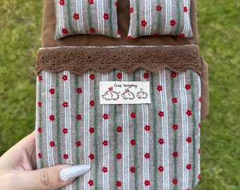 May include: A miniature bed set featuring a brown headboard and two matching pillows with a grey and white striped pattern accented by small red flowers. The bed cover has a brown lace trim and a white label with the text Cute hedgehog.