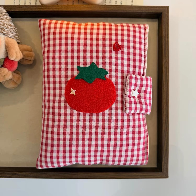 May include: A rectangular fabric notebook cover featuring a red and white gingham pattern. The cover is decorated with a textured red tomato applique with a green stem, a small red heart button, and a white star button on the side closure tab.
