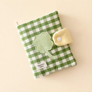 May include: A fabric-covered notebook featuring a green and white gingham pattern. The cover is decorated with a light green fabric clover appliqué and a small white label reading Cotton Diary. A pale yellow tab with a white snap closure secures it.
