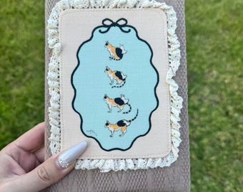 May include: A rectangular notebook with a textured beige cover, framed by white lace trim. The centre features a light blue scalloped panel with a black bow graphic at the top and four illustrations of a calico cat in various poses.