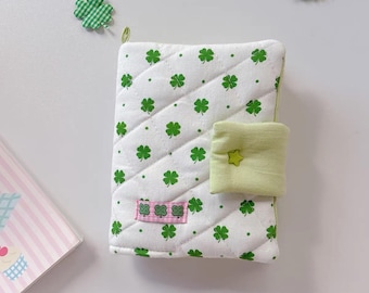 May include: A white quilted notebook cover featuring a green shamrock design. The cover has a light green tab closure with a star-shaped button. A pink and green label with shamrocks is sewn on the front. Shamrock accents are scattered around the cover.