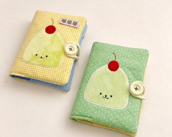 May include: Two fabric-covered notebooks featuring plush, light green character appliques with red cherry tops. One notebook has a yellow gingham cover, the other a green polka-dot cover. Both include a white button closure with a cord loop.