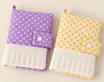May include: Two quilted fabric notebook covers, one purple with white polka dots and one yellow with purple polka dots. Both feature a white lace trim, a vertical quilted bottom section, and a side strap with a white snap button closure.