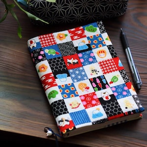 May include: A colorful book cover with a patchwork design featuring various Japanese-inspired motifs. The cover includes squares of red, white, blue, and black fabric with illustrations of cats, flowers, and other symbols. A black pen rests beside the book.