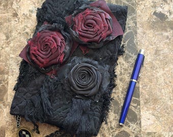 May include: A black fabric journal cover featuring three dimensional fabric roses in deep burgundy and black, with frayed edges and lace trim. A small oval charm with a black and white checkered pattern hangs from the corner, accompanied by a blue pen.
