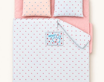 May include: A miniature bedding set featuring a solid pink base, two rectangular pillows with polka dot patterns, and a white blanket with red polka dots. A small rectangular patch on the blanket displays a blue rabbit graphic and the text Let's go.