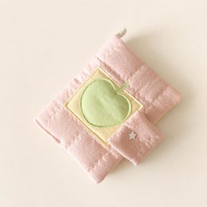 May include: A quilted pink fabric pouch featuring a square patch with a light green apple appliqu&eacute;. The pouch includes a smaller attached pocket adorned with a tiny white star-shaped embellishment, secured with a small fabric loop at the top corner.