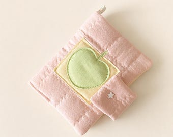 May include: A quilted pink fabric pouch featuring a light yellow square patch with a centred light green apple appliqué. A smaller matching pink flap is attached to the front, adorned with a small white star-shaped button detail.