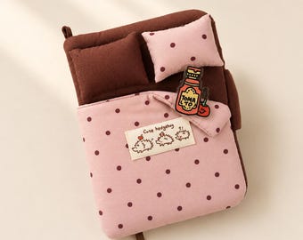 May include: A plush bed-shaped organiser featuring a dark brown base, a solid dark brown pillow, and a light pink pillow with dark brown polka dots. A light pink blanket with dark brown polka dots covers the bed, displaying a label with the text "Cute hedgehog" and a small acrylic charm shaped like a bottle labelled "TOMA TO" with a tomato illustration.
